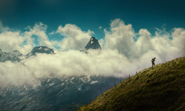 A hiker walking uphill on a grassy slope with mountains and clouds in the background