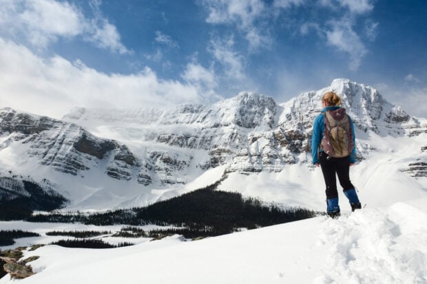 A hiker wearing a backpack stands on snowy terrain facing a mountain range covered in snow under a blue sky