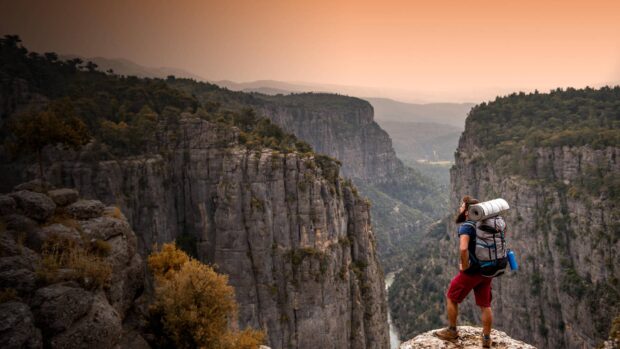 A man with hiking gear overlooking a deep canyon with lush forest and rocky cliffs at sunset