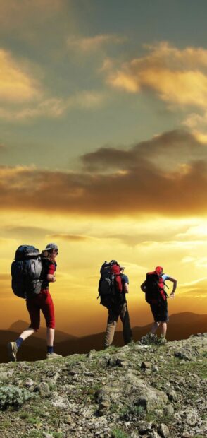 Three hikers wearing backpacks trekking on a rocky trail during sunset with a colorful sky