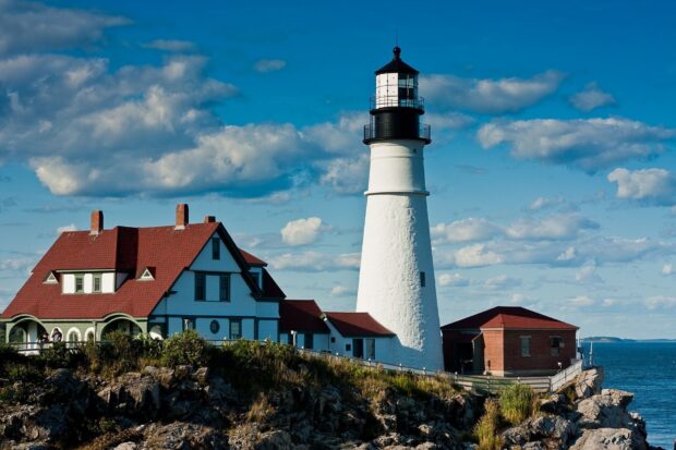 A historic Maine lighthouse stands tall on a rocky coast with a blue sky in the background, HD Desktop Wallpaper
