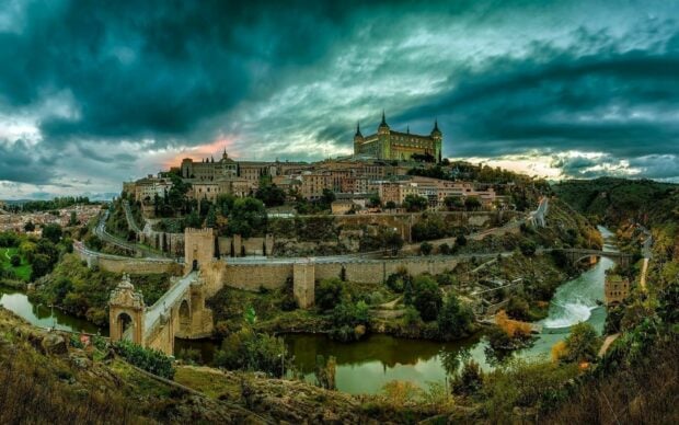 A scenic view of a historic Spanish city with a river flowing around ancient walls under a dramatic sky
