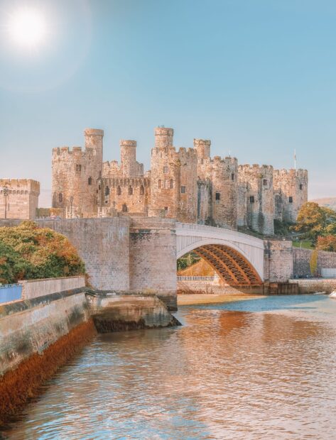 A historic stone castle stands beside a calm river under a bright blue sky in Wales, HD Desktop Wallpaper