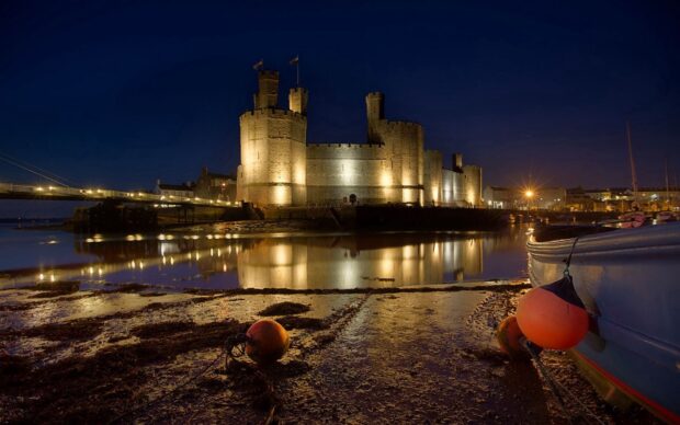 A historic castle in Wales is illuminated against a dark night sky with reflections in the water, HD Desktop Wallpaper