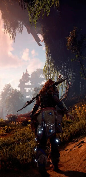 Aloy exploring a lush forest path with towering ruins visible in the distance