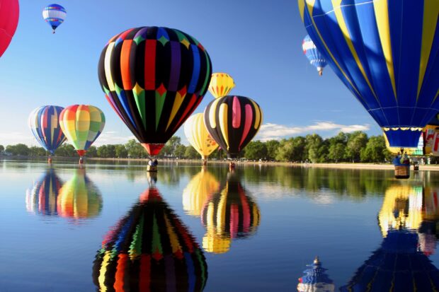 Colorful hot air balloon flying over a calm lake with clear sky and trees in the background