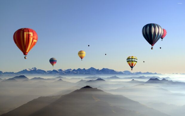 Colorful hot air balloons floating over misty mountains during sunrise with clear blue sky