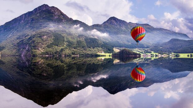 A colorful hot air balloon floating over a calm lake reflecting mountains and sky
