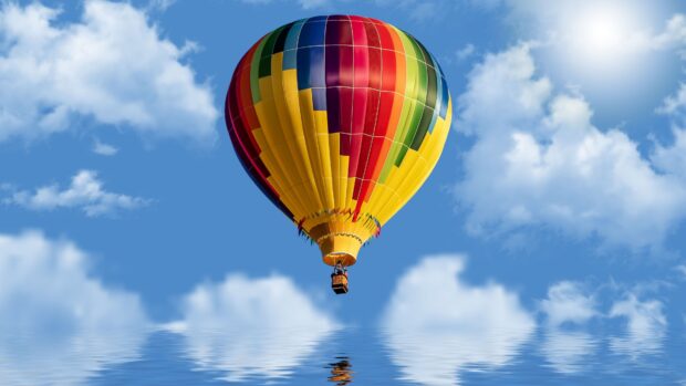 A colorful hot air balloon floating above a calm reflective water surface with blue sky