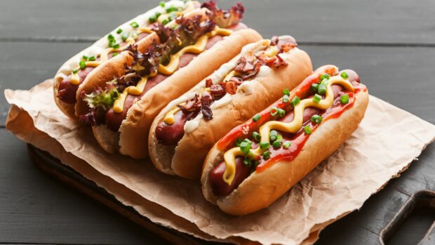 Four hot dog with various toppings including mustard lettuce bacon and chives on parchment paper