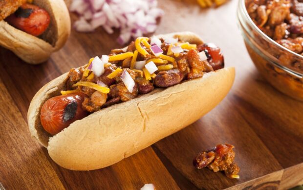 A chili topped hot dog with chopped onions and shredded cheese on a wooden surface