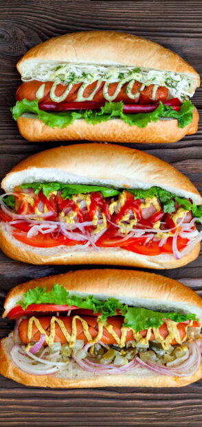 Three hot dog sandwiches with various toppings and fresh vegetables on wooden surface