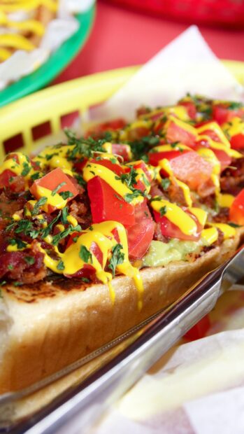 Hot dog topped with fresh tomatoes and mustard drizzled on a toasted bun