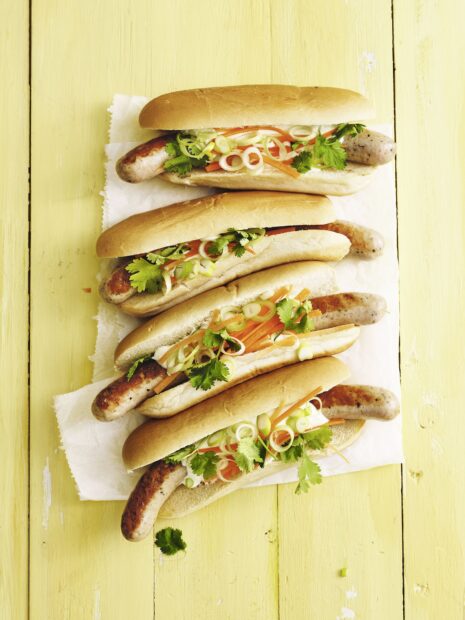 Four hot dog sandwiches with fresh herbs and vegetables on a yellow wooden surface