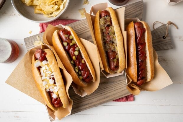 Four hot dogs with different toppings served on a wooden board with chips in the background