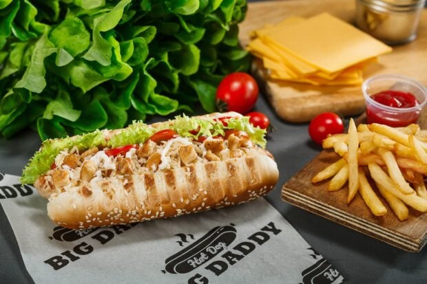 Grilled hot dog with chicken pieces and fresh vegetables on a sesame bun served with fries and cheese