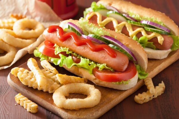 Hot dog with fresh vegetables and ketchup served with fries and onion rings on wooden board