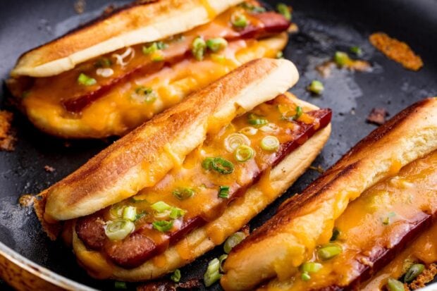 Hot dog with melted cheese and green onions in a toasted bun on a frying pan