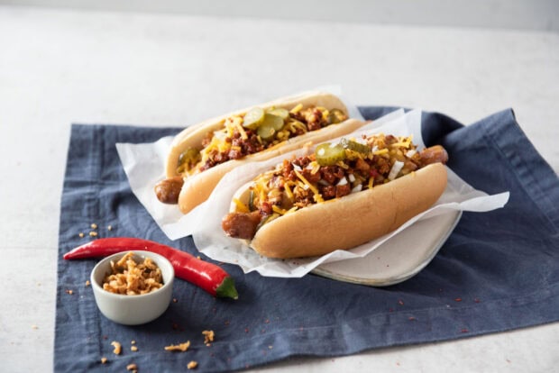 Two hot dog with toppings and chili pepper on a white plate