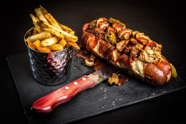 A hot dog with pulled meat and sauce served with seasoned fries on a black slate