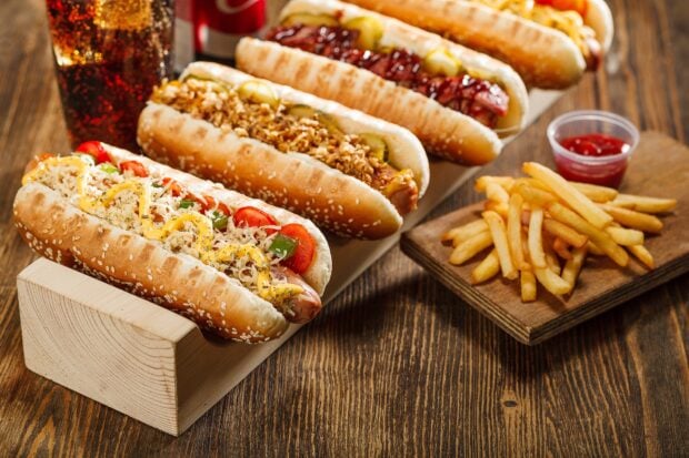A variety of hot dog with different toppings served on a wooden rack alongside fries and a glass of soda