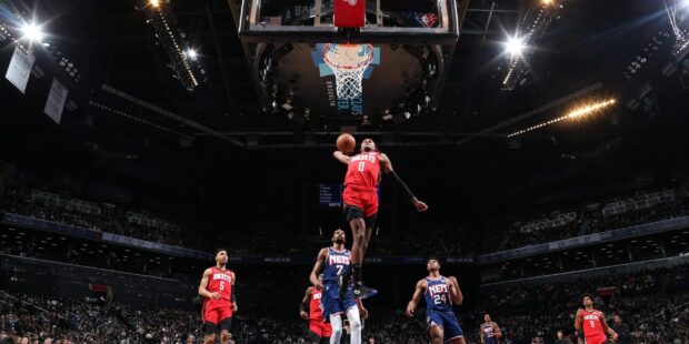 A Houston Rockets player soaring for a dunk during a basketball game against the Nets in a crowded arena