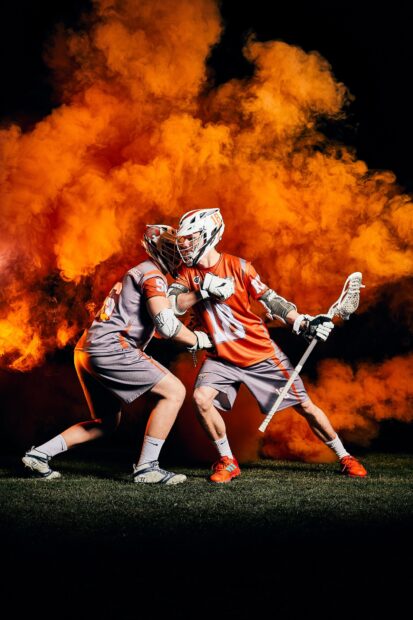 Two players in lacrosse gear compete fiercely with vivid orange smoke behind, HD Phone Wallpaper
