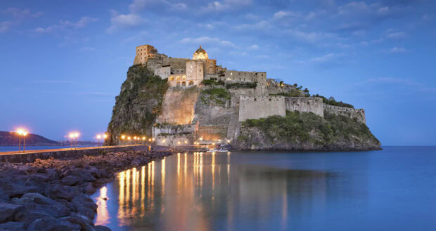 Historic castle on rocky island in Ischia during twilight with calm sea and lights reflection