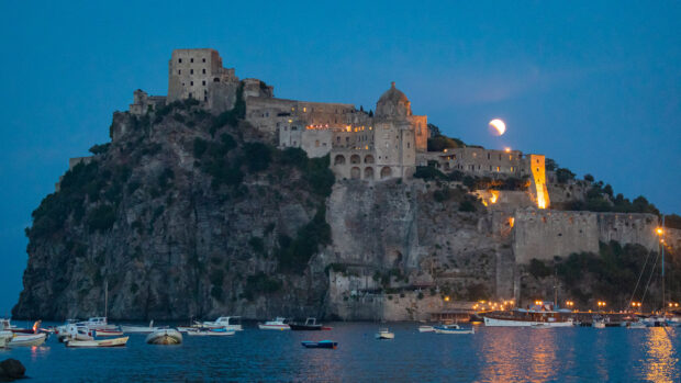 Ischia fortress lit up at dusk with boats in the water under a moonlit sky