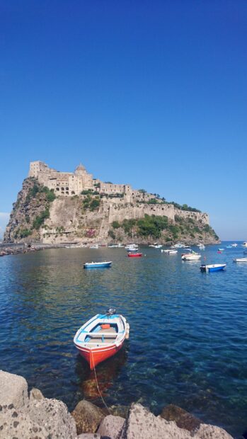 A fishing boat floating on clear water near a rocky coast with Ischia castle in the background