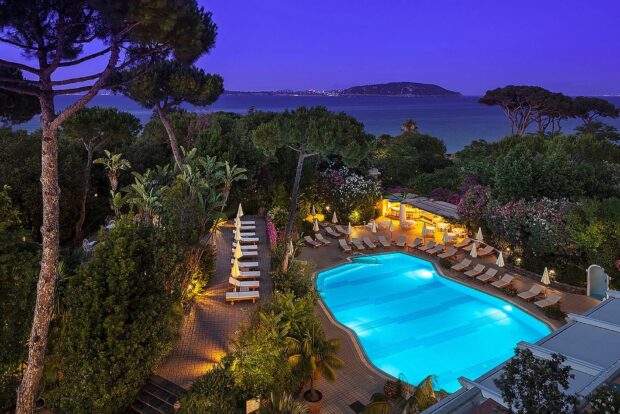 A scenic view of Ischia with a lit swimming pool surrounded by trees and ocean in the background