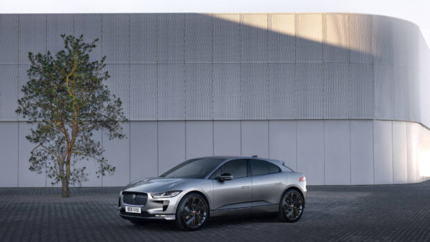 A silver Jaguar I Pace is parked near a modern building and tree, 2K Desktop Wallpaper