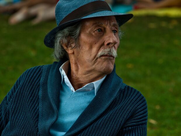 An elderly man wearing a blue hat and sweater looks to the side thoughtfully in an outdoor setting, HD Desktop Wallpaper