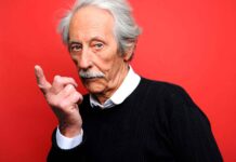 Senior man with white hair and mustache making a shushing gesture against red background.