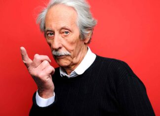Senior man with white hair and mustache making a shushing gesture against red background.
