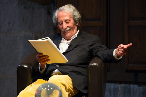 Jean Rochefort is sitting and smiling while reading a script on stage, HD Desktop Wallpaper