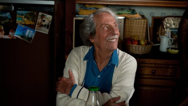 An elderly man wearing a blue shirt and white sweater is smiling warmly in a cozy home setting with a basket of fruit in the background, 4K Desktop Wallpaper