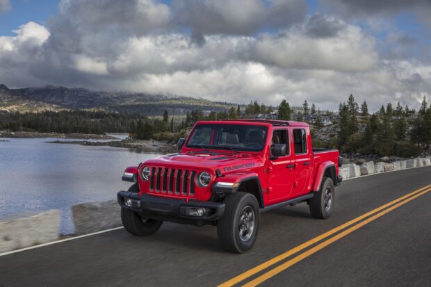A red Jeep Gladiator driving on a mountain road beside a lake under cloudy skies