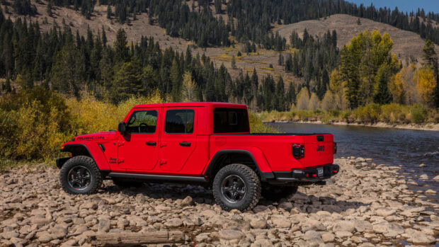 Red Jeep Gladiator parked on rocky riverbank in forested mountain area