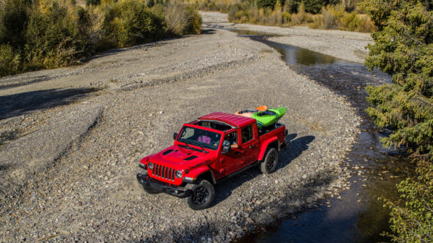 Red Jeep Gladiator carrying kayaks on a rocky riverbed surrounded by trees