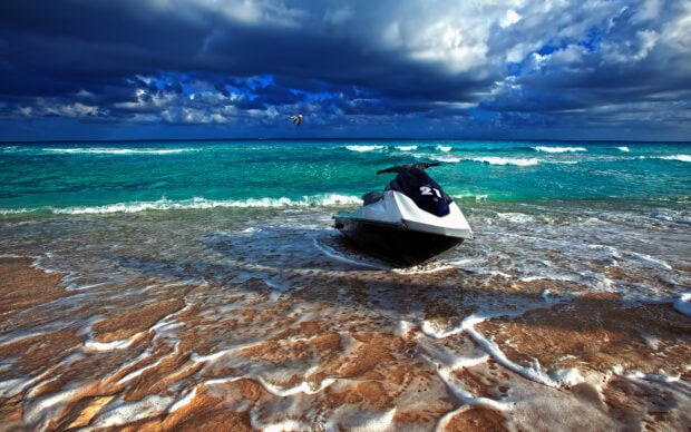 A jet ski rests on the sandy shore with turquoise ocean waves and a cloudy sky in the background, HD Desktop Wallpaper