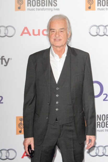 Jimmy Page wearing a black suit and white shirt standing against a branded event backdrop