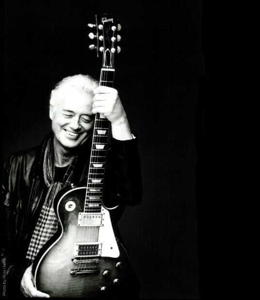 Jimmy Page holding a Gibson guitar with a joyful expression on a dark background