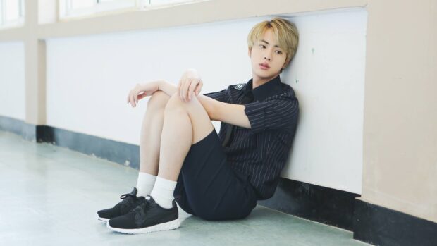Jin BTS sitting indoor with a thoughtful expression near a white wall, HD Desktop Wallpaper