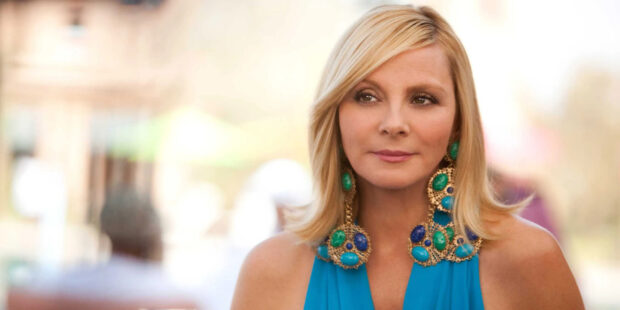 Kim Cattrall wears a blue dress and large earrings in a calm outdoor setting, 2K Desktop Wallpaper
