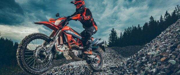 Ktm dirt bike rider performs off road stunt against a cloudy sky and rocky terrain background