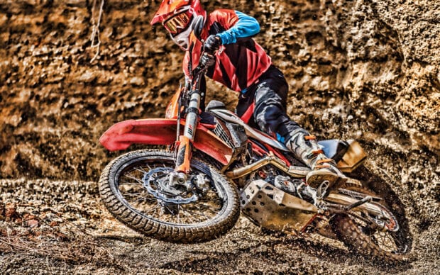 A rider in full gear aggressively turns a KTM dirt bike on a rugged dirt trail