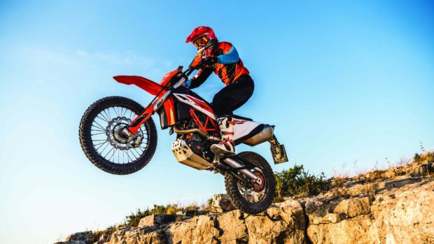 A rider jumps a KTM dirt bike over rocky terrain under a clear blue sky