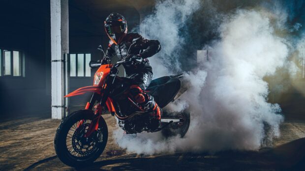 A rider performs a burnout on a KTM dirt bike creating thick smoke inside a dimly lit garage