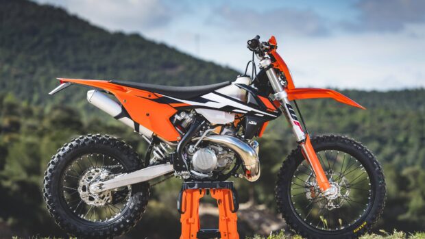 A bright orange KTM dirt bike is displayed outdoors with a mountain background